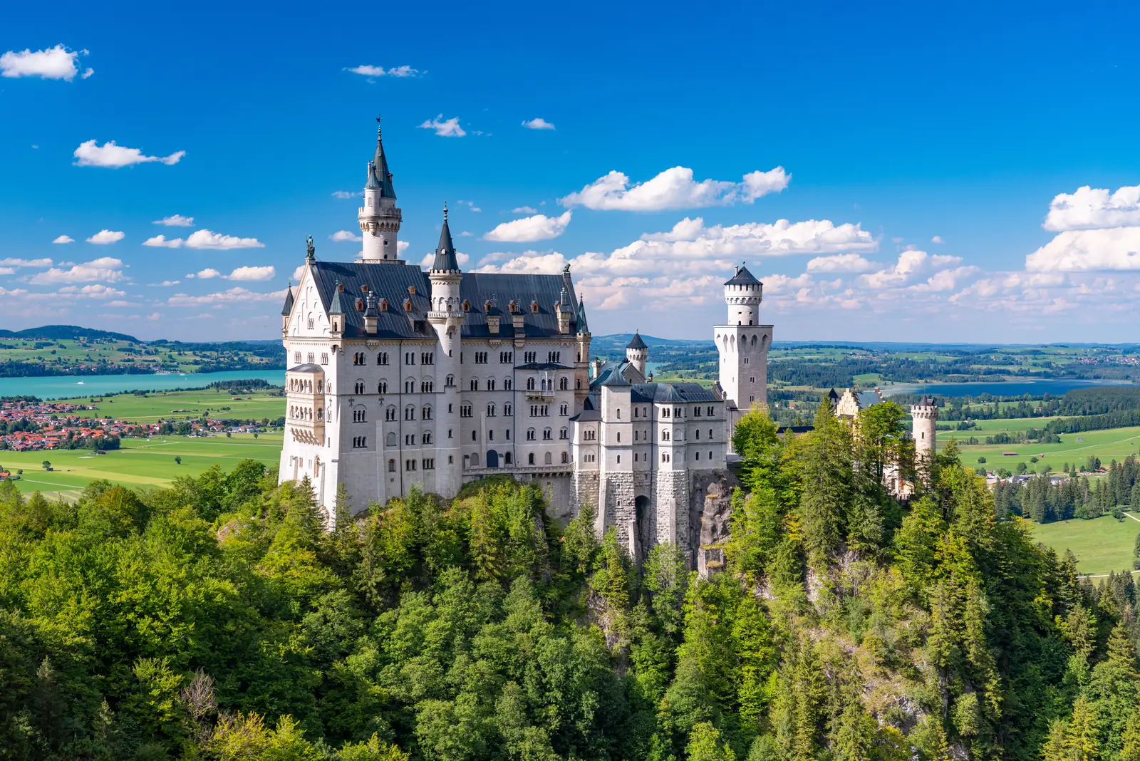 El castillo blanco de Neuschwanstein sobre una colina boscosa, con campos y un lago al fondo.
