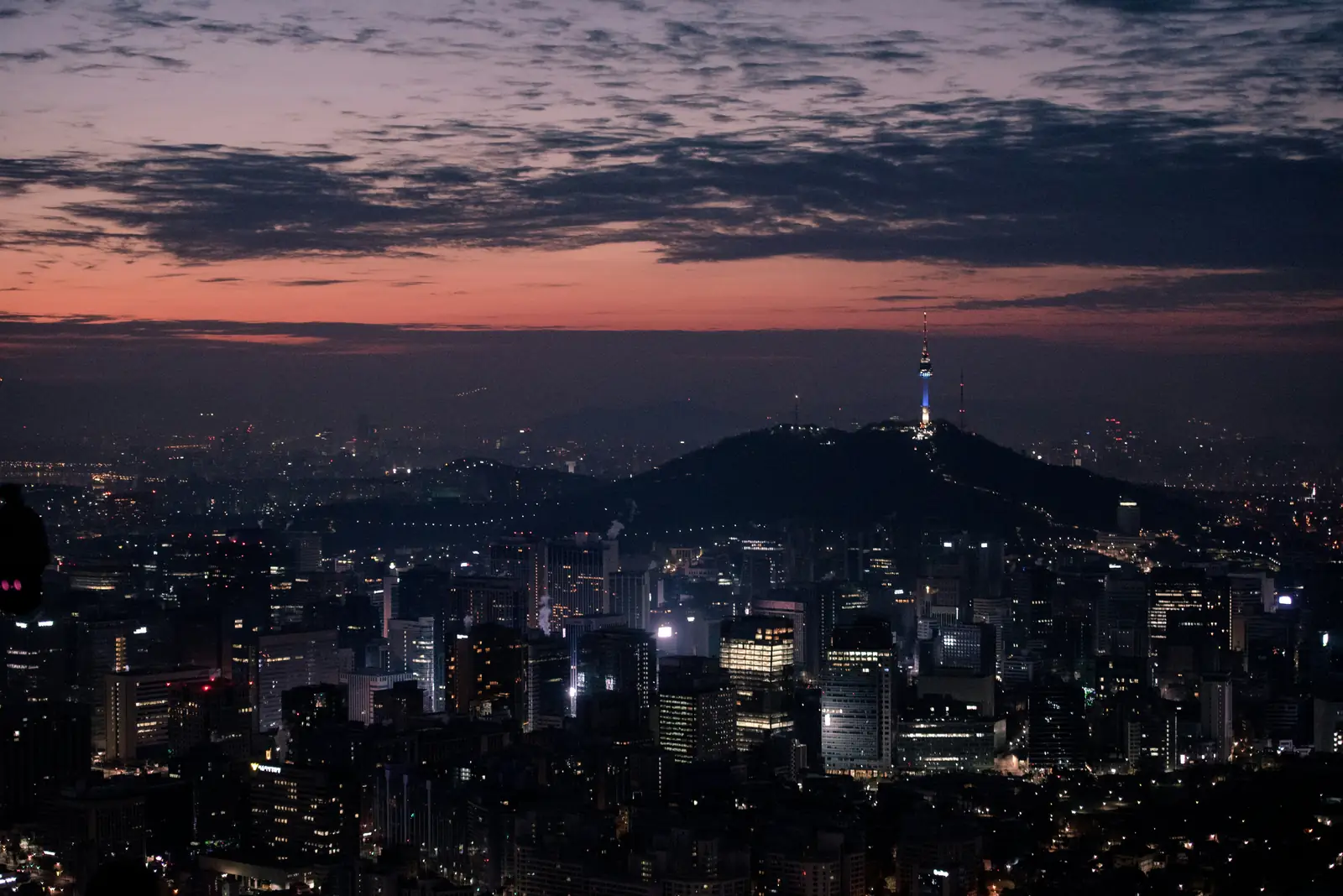 Vista nocturna de Seúl desde lo alto, con la torre Namsan iluminada sobre una colina y un cielo de atardecer rojizo.