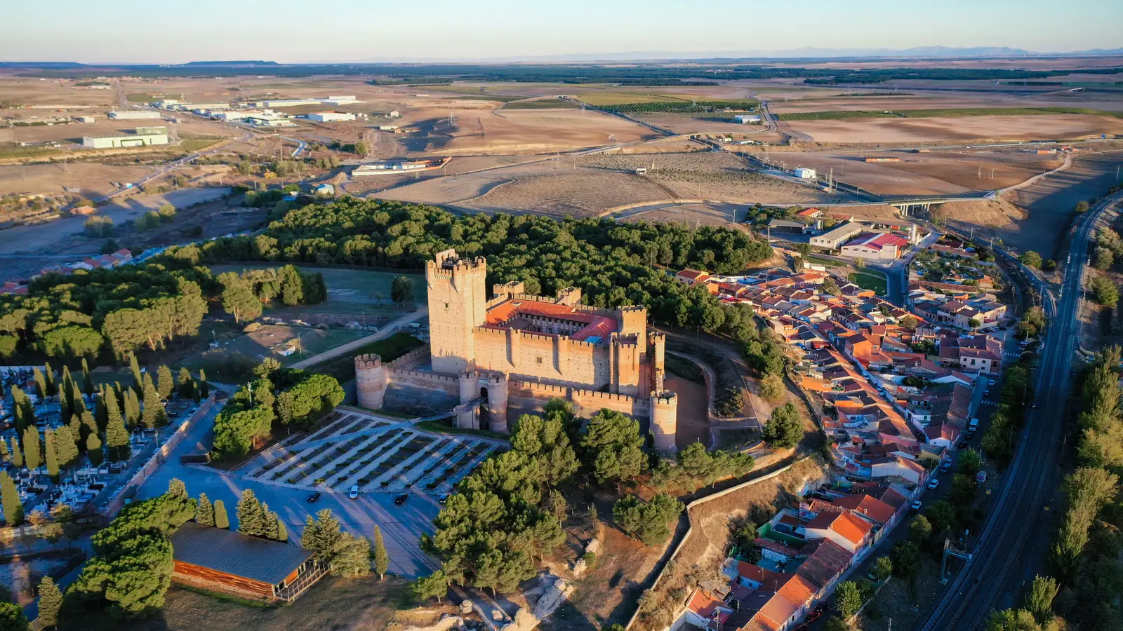 Vista aérea de un castillo de ladrillo rodeado de pinos, con el pueblo y los campos de Castilla alrededor.