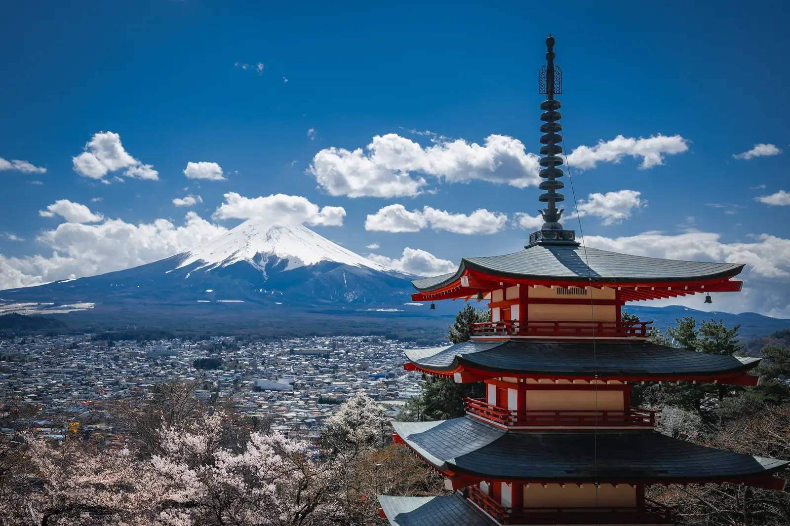 La pagoda Chureito en primer plano, con el monte Fuji nevado y la ciudad de Fujikawaguchiko al fondo, bajo un cielo azul con nubes.