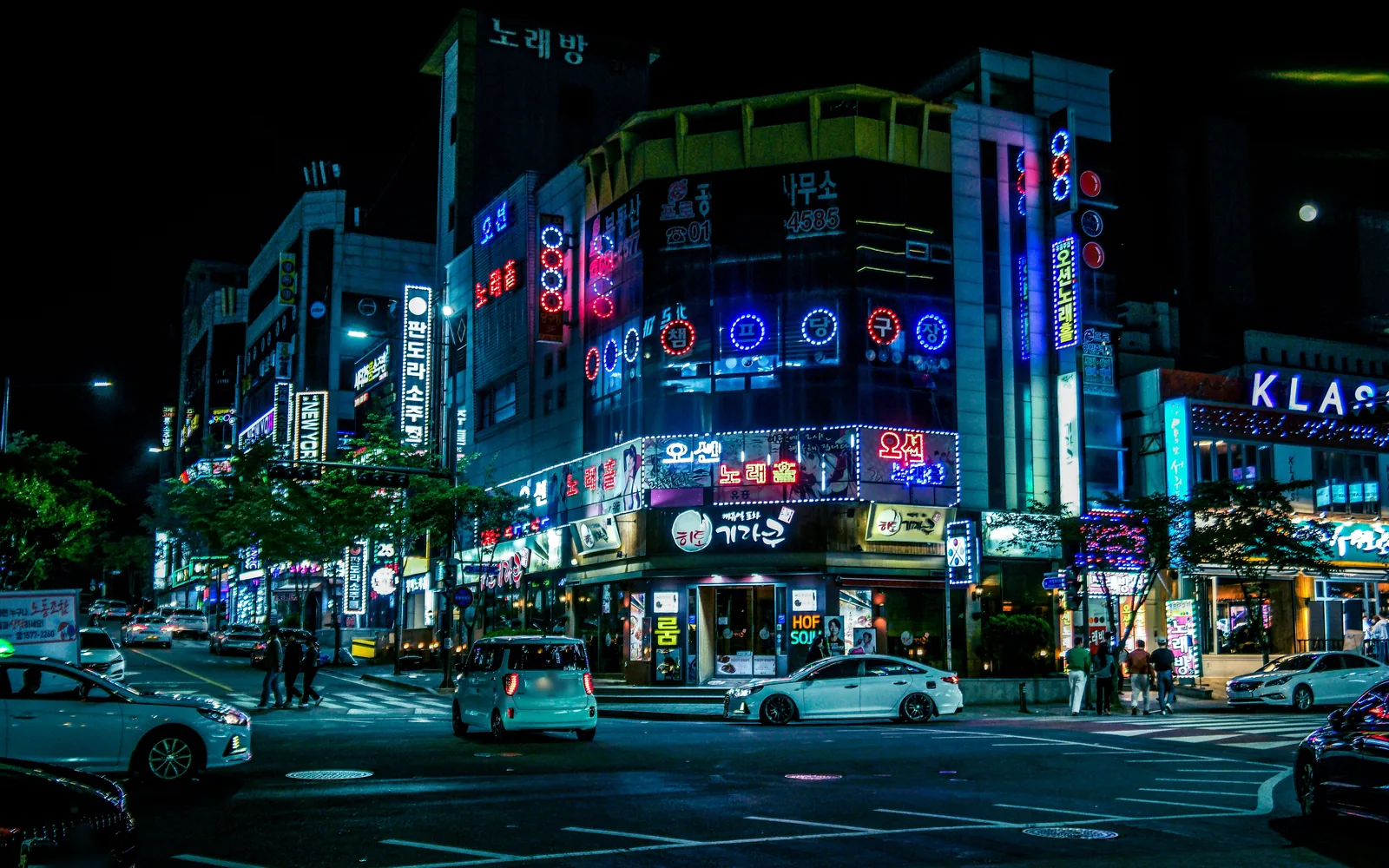 Una calle de noche con letreros de neón de colores en un edificio de esquina, y coches en el cruce.