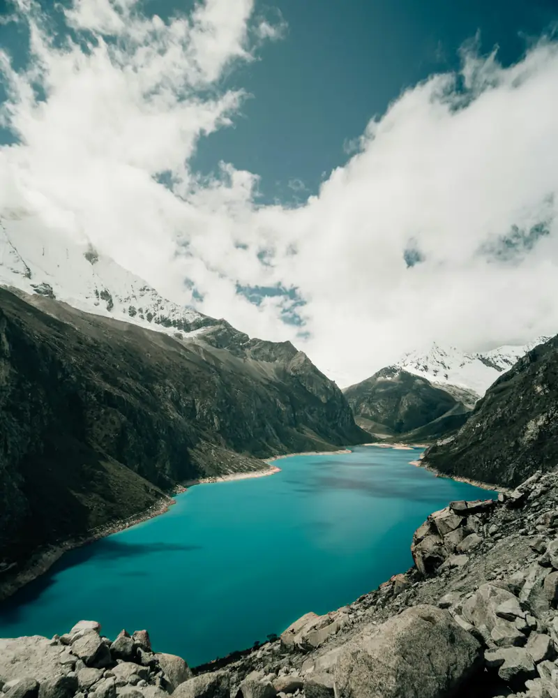 Una laguna de agua turquesa entre montañas rocosas y picos nevados, bajo nubes.
